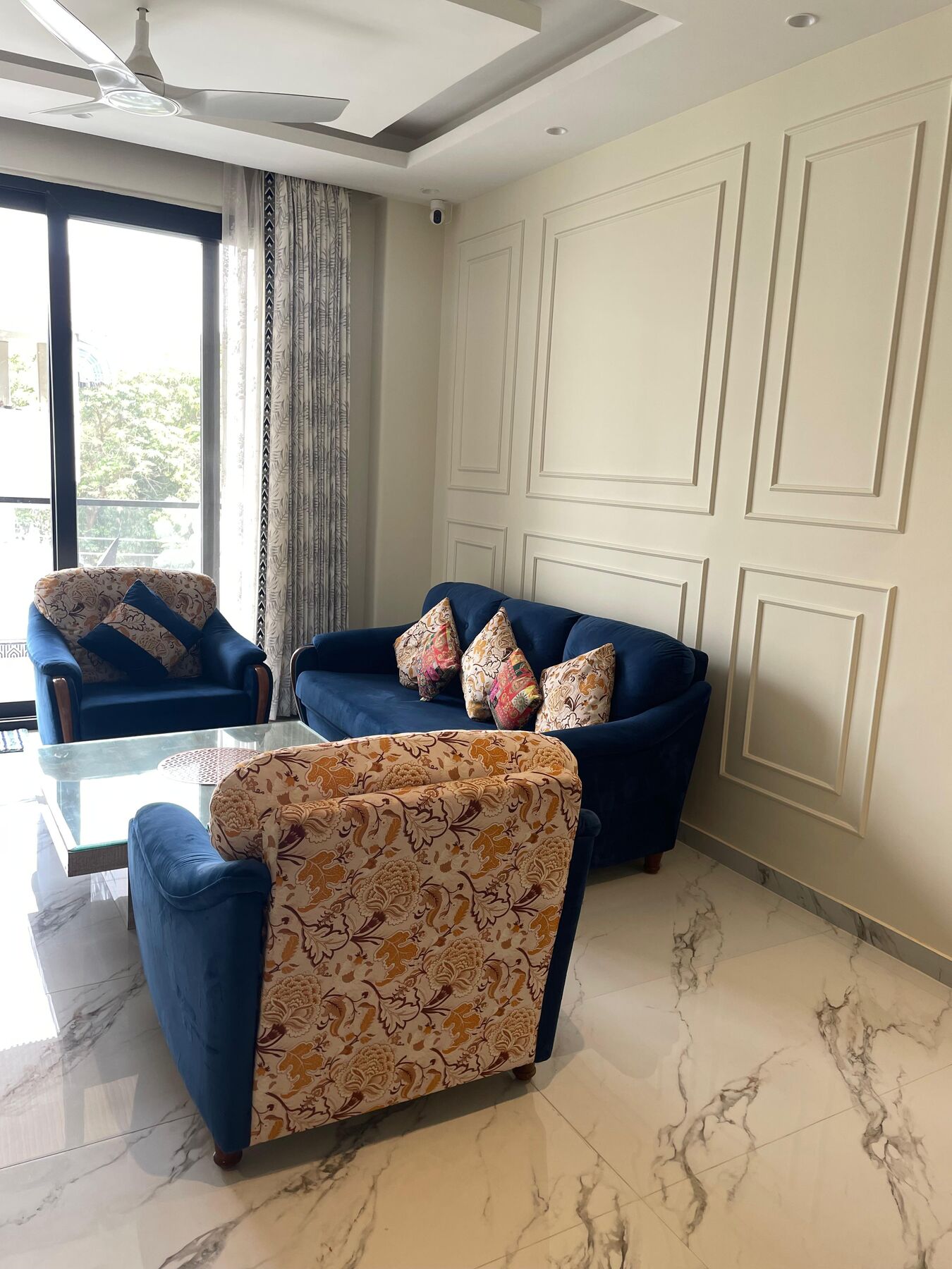 The living room as handed over: deep blue velvet sofas and a floral
                  armchair drawn round a glass table, against the panelled wall and the
                  marble floor.