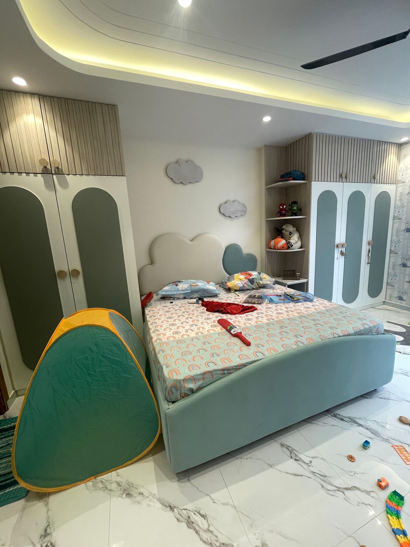 A child's bedroom: a teal upholstered bed under cloud-shaped joinery, a
                  rounded chair beside it and the wardrobes in pale green behind.