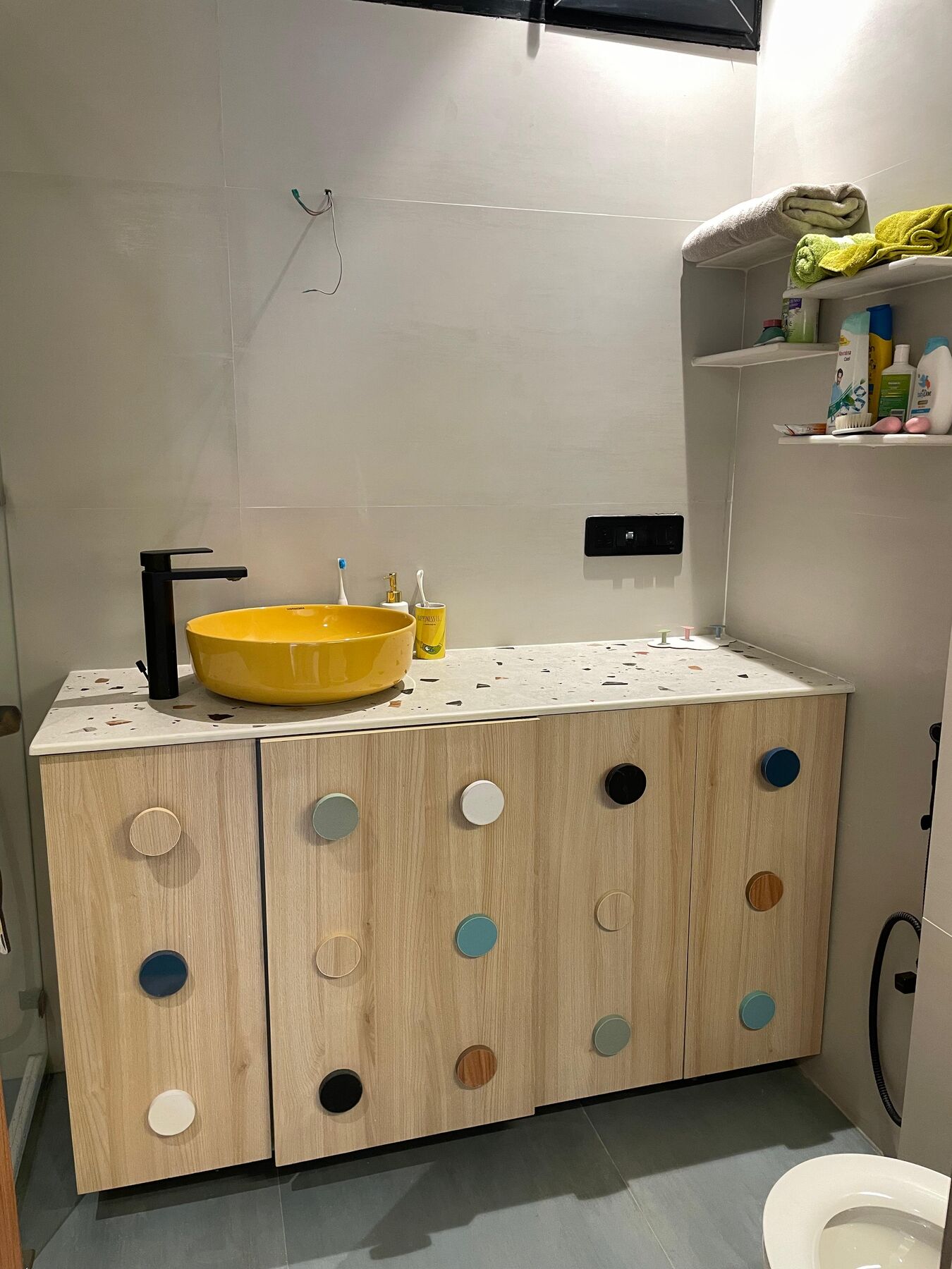 A bathroom: a yellow basin set on a timber vanity dotted with coloured
                  discs, the tap and shelving above it in black.