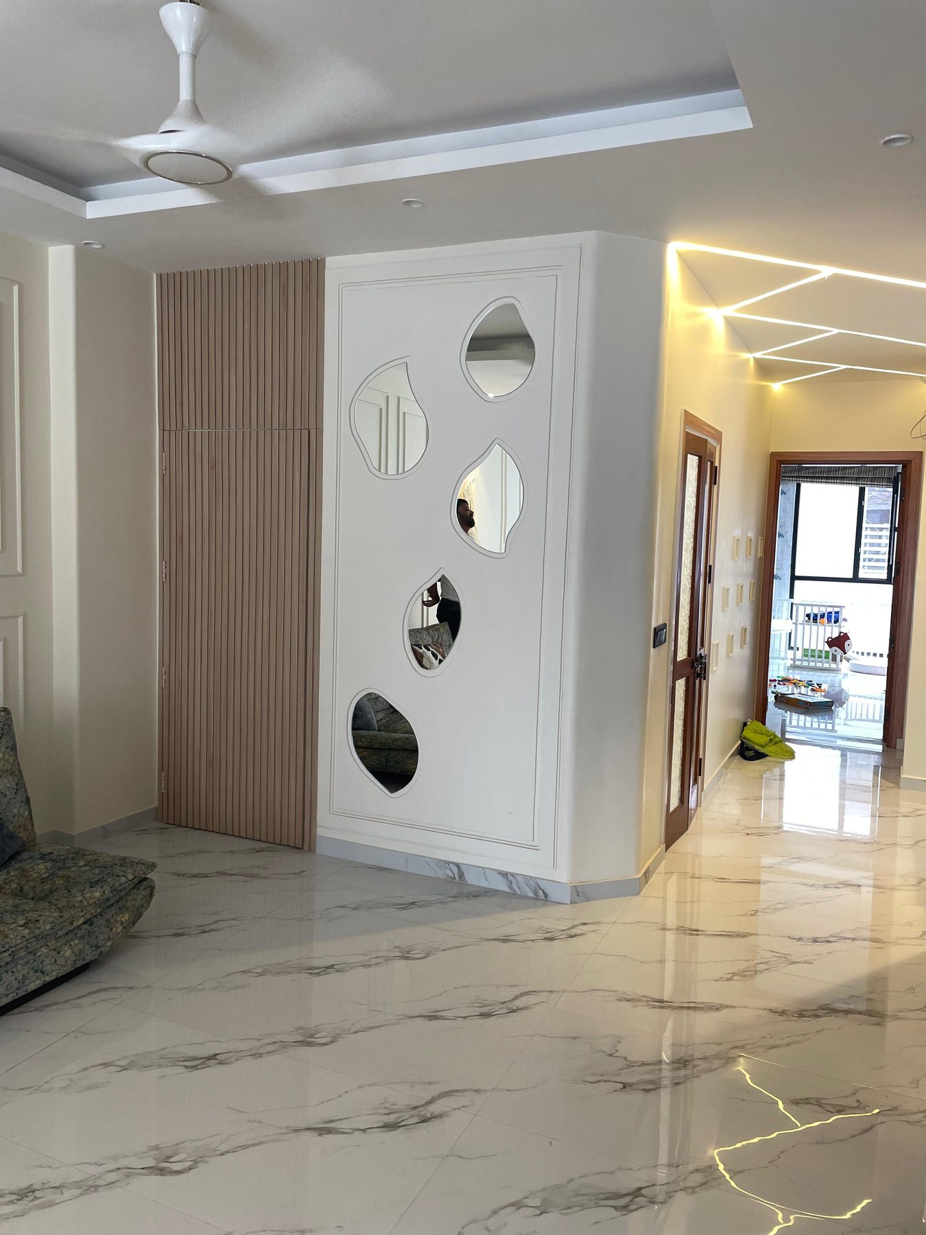 A partition set with organic mirror cut-outs of varying sizes, beside
                  a fluted timber panel, dividing the corridor from the living room.