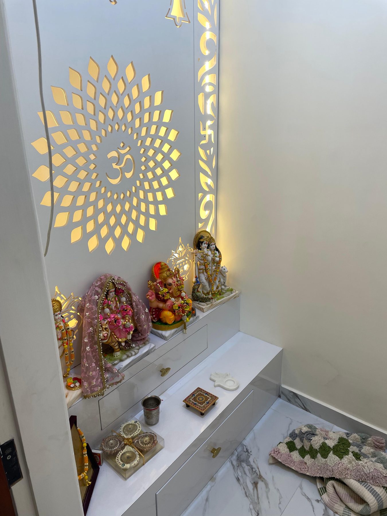 The pooja: a gold Om sunburst cut into a backlit panel above a white
                  marble ledge holding the idols.