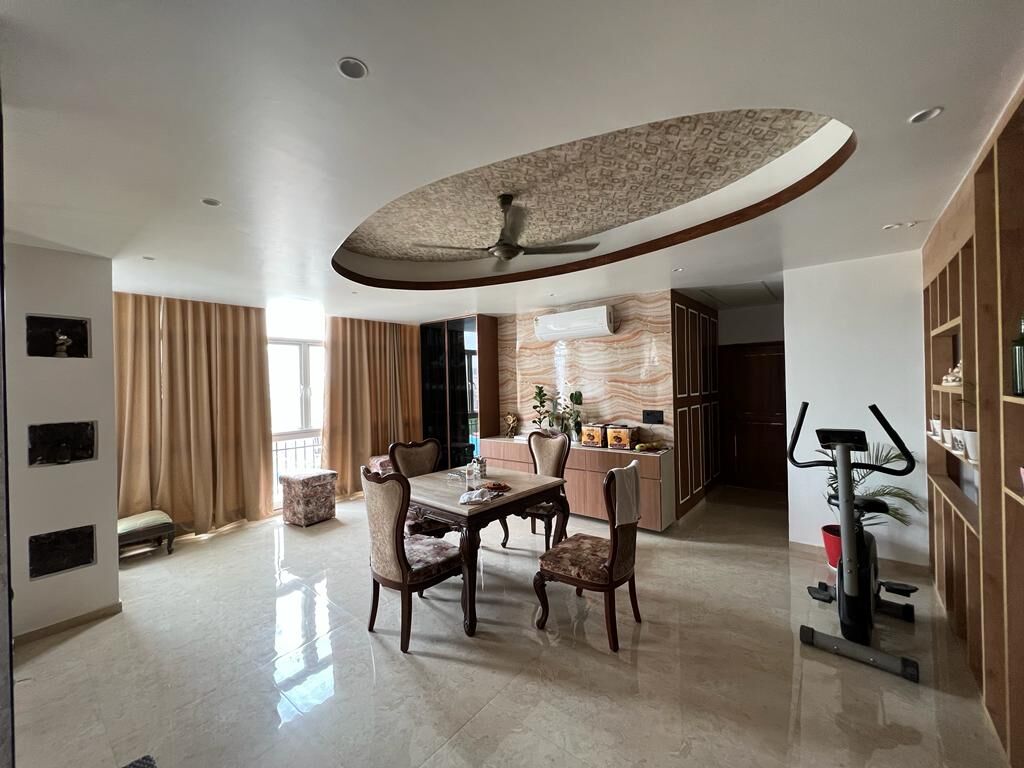 The dining end: an oval timber raft in the ceiling with a fan below it,
                  carved chairs drawn up to the table, and the exercise corner past the
                  curtains.